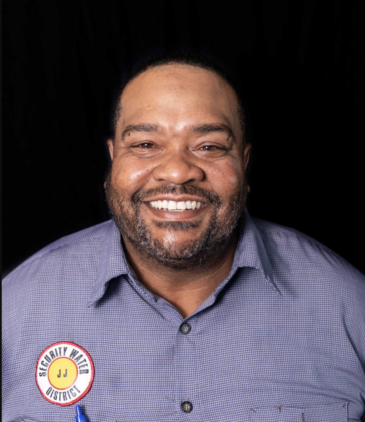 Headshot of JJ Campbell in a blue button-down security water shirt, smiling in front of a dark background